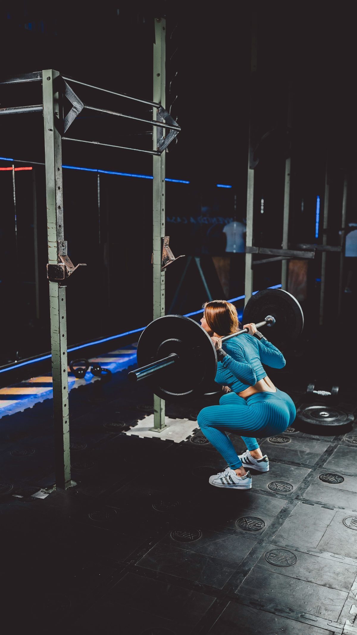 Kobieta w turkusowym stroju sportowym wykonuje przysiad ze sztangą na plecach przy klatce treningowej