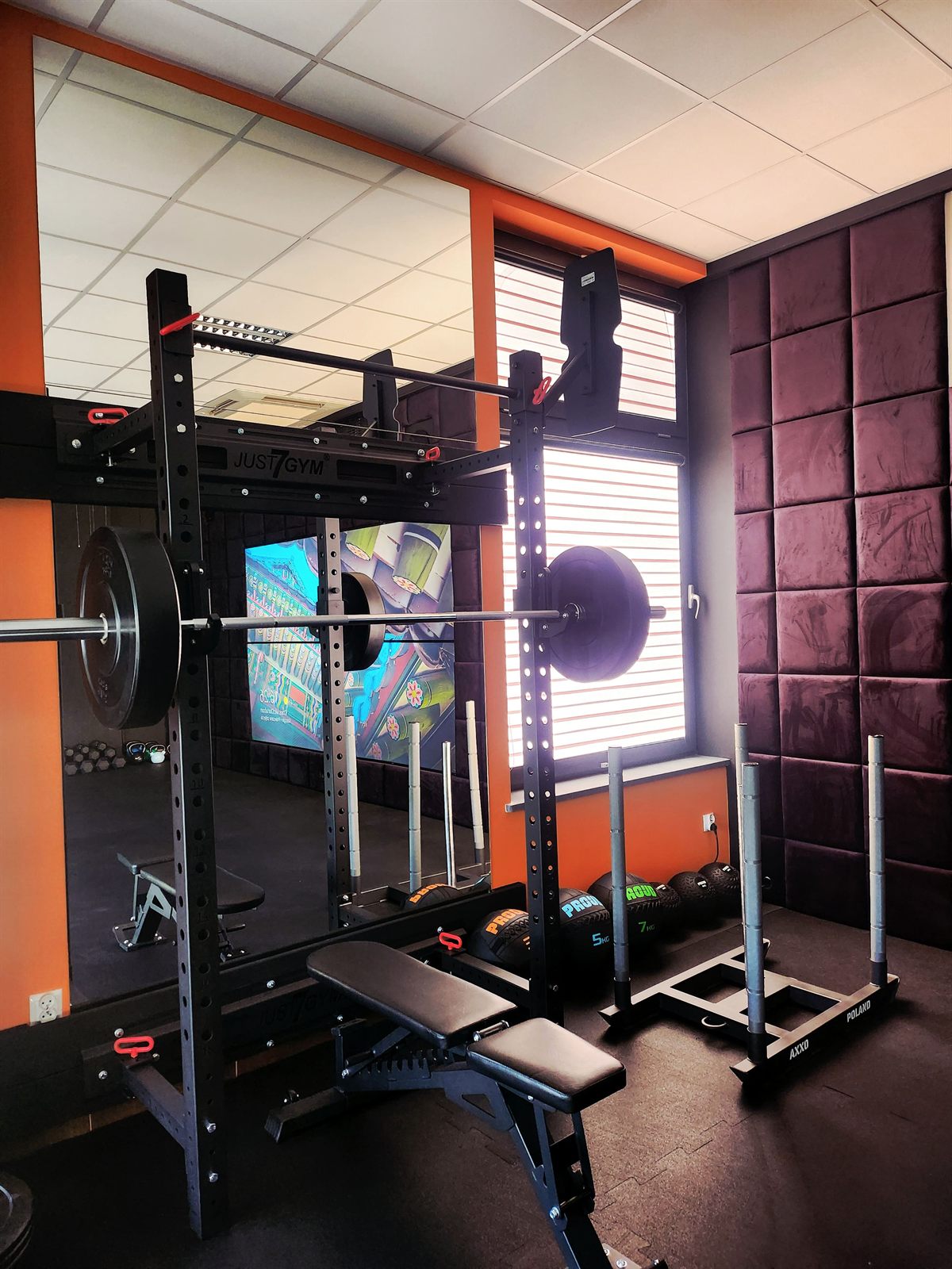 Strefa siłowa — klatka treningowa Just7Gym ze sztangą i talerzami, ławka regulowana, piłki lekarskie i sanie treningowe przy oknie